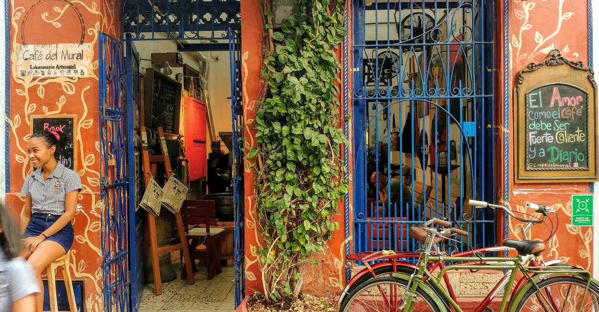 Fachada de Café del Mural en Getsemaní: muro terracota con murales pintados a mano, rejas azul cobalto, plantas colgantes, bicicleta roja y un tablero de tiza con la frase El Amor como el Café debe ser Fuerte, Caliente y a Diario.